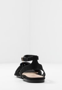 Sandale en daim noir avec un design enroulé sur le devant, bride de cheville avec quincaillerie dorée, semelle plate et talon ouvert. Forme minimaliste et contemporaine.
