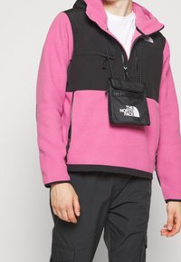 Chaqueta de forro polar rosa con una sección superior negra, bolsillos con cremallera y un pequeño pouch negro con un logotipo. Presenta capucha y cierre de cremallera.
