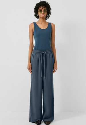 Vrouw met krullend haar, groene zonnebril, een blauw mouwloos topje, wijde broek met trekkoord en zwarte schoenen, staand tegen een grijze achtergrond.