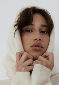 Biała bluza z kapturem o miękkiej fakturze, model trzymający materiał blisko twarzy, naturalne tony skóry i neutralne tło.