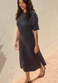 Robe midi gris foncé à manches courtes, texture douce, fente sur le côté et coupe décontractée. Associée à des sandales simples, sur un fond clair.