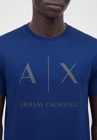Άτομο που φοράει μπλε T-shirt Armani Exchange με μεγάλο γκρι κείμενο "A | X" και "ARMANI EXCHANGE" στο μπροστά μέρος.