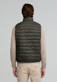 Vesta puffer de culoare verde măsliniu, cu quilting orizontal, guler înalt. Purtați-o peste un pulover bej, asortată cu pantaloni deschiși. Textură netedă și lucioasă.