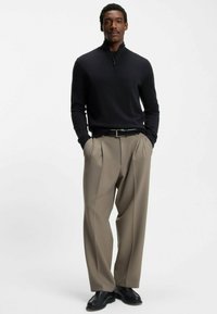 Pull noir à col zippé en tissu doux, associé à un pantalon beige plissé. Le modèle porte des chaussures en cuir noir, debout devant un fond uni.