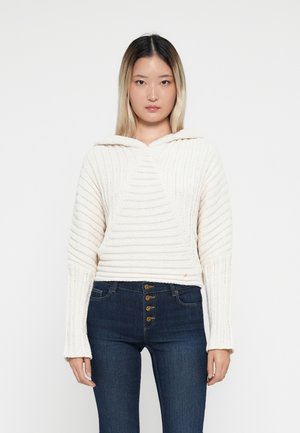 Kvinna som står mot en vit bakgrund iförd en krämfärgad stickad huvtröja och mörkblå hög midja jeans med knapplåsning.