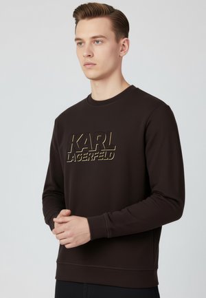 Junger Mann mit ordentlich gestyltem Haar, trägt ein dunkelbraunes Langarmshirt mit dem "KARL LAGERFELD"-Logo auf der Brust, die Hände ineinander verschränkt.
