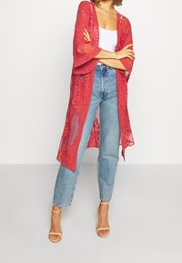 Punainen pitsineuletakki kukkakuoseilla, avoin edessä, kolme neljäsosankaan hihat, valkoisen topin päällä ja sinisten farkkujen kanssa yhdistettynä.