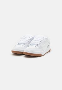 Zapatillas blancas con parte superior de cuero suave y textil, diseño de cordones, cuello acolchado y suela de goma marrón. Sin patrones ni acentos visibles.