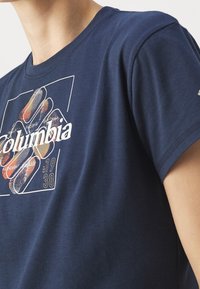 Tmavě modré bavlněné tričko s grafickým motivem, který obsahuje nápis "Columbia" a šestíhranný vzor v tlumených barvách. Krátké rukávy.