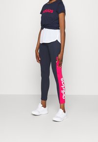 Mörkblå t-shirt med rosa "adidas"-logotyp, vit lagerkant; mörkblå leggings med rosa sidpanel och vit "adidas"-text; vita sportskor.
