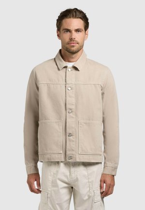 Hombre con chaqueta beige abotonada con grandes bolsillos delanteros y pantalones cargo crema, de pie contra un fondo claro y sencillo.