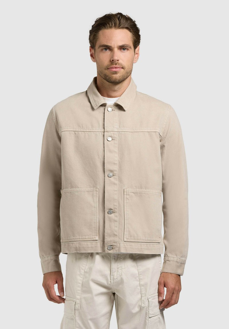 Homme portant une veste beige à boutons avec de grandes poches avant et un pantalon cargo crème, debout devant un fond clair uni.