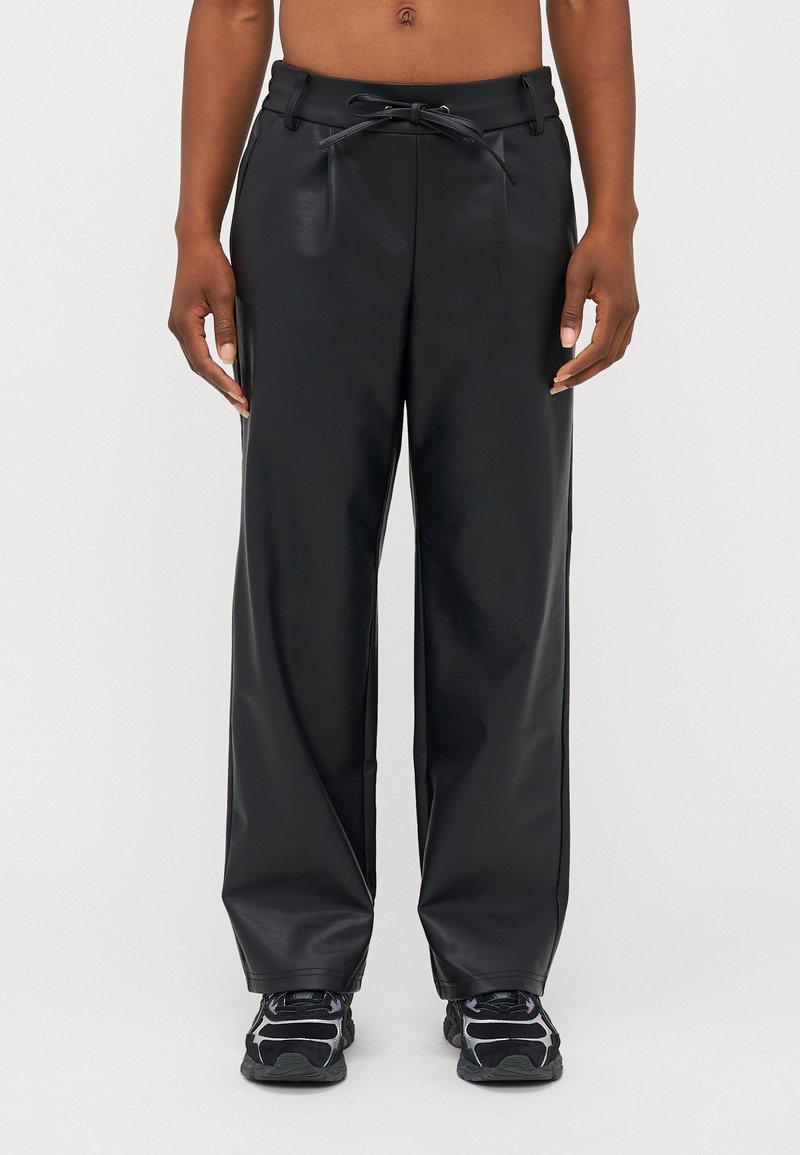 Zwarte nep leren wijde broek met een trekkoord in de taille, voorzien van zijzakken en een gladde textuur. G gedragen met zwarte sneakers.