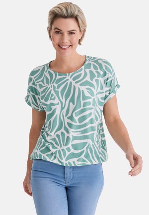 Lächelnde Frau mit kurzen blonden Haaren, die ein hellgrünes und weiß gemustertes kurzärmeliges Oberteil und hellblaue Jeans vor weißem Hintergrund trägt.