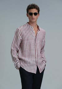 Licht gestreepte shirt in rood en wit, dichtgeknoopt met een bandkraag, lange mouwen en een relaxte pasvorm. Gecombineerd met zwarte broek. Zonnebril op.