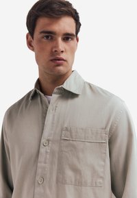 Lichtgrijze button-up shirt gemaakt van textuurstof. Kenmerkt zich door een puntige kraag en een enkele borstzak.