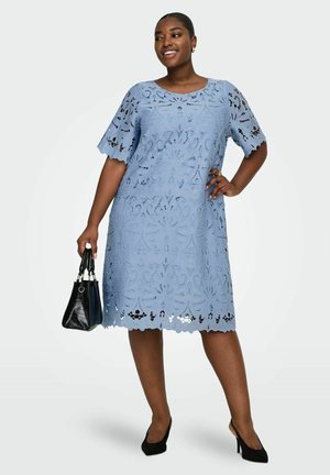 Femeie purtând o rochie albastră de dantelă, până la genunchi, ținând o geantă neagră și încălțând tocuri negre, zâmbind cu o mână pe șold.