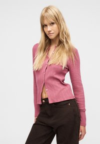 Femme blonde portant un cardigan rose boutonné et un pantalon brun foncé, debout avec une main près de sa poche contre un fond uni.