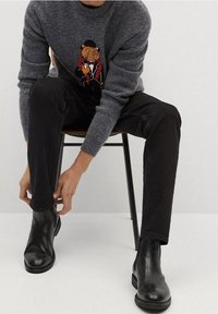 Pull en laine gris avec un motif de tigre brodé, associé à un pantalon en denim noir et des bottines en cuir noires, assis sur une chaise en bois.