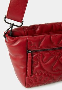 Červená kožená kabelka přes rameno s vyraženými květinovými vzory, obsahující zipovou kapsu, polstrovaný design a nastavitelné ramenní popruhy s kovovými prvky.