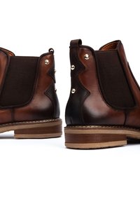 Bottes Chelsea en cuir marron avec panneaux élastiques, clous décoratifs et semelle texturée. Talon bas empilé et bout arrondi complètent le design.