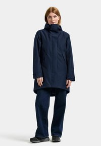 Marinblå vattentät jacka med hög krage och huva, med sidofickor och knälång fåll. Bärs med mörkblå vida jeans och skor.