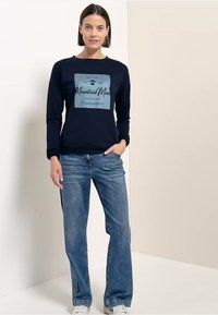Frau in einem marineblauen Sweatshirt mit dem Aufdruck "Mountain Moon" und blauen Jeans, steht mit einer Hand in der Tasche vor einem einfarbigen Hintergrund.