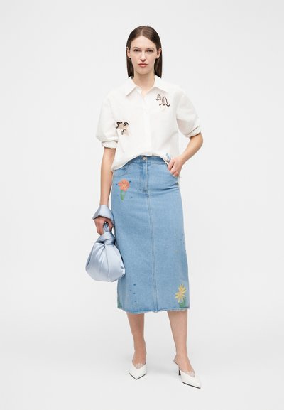 Kvinne iført en hvit skjorte med soppbroderi, denimskjørt med blomsterbroderi, hvite hæler, og holder en blå satengveske.