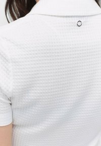 Vit texturerad polo med ett våffelmönster, som har en rundad krage och en liten svart cirkulär detalj på baksidan.