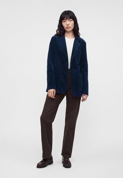 Femme portant un blazer bleu marine, un haut blanc, un pantalon marron et des mocassins marron foncé, debout devant un fond gris clair.