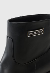 Černé kožené kotníkové boty s hladkým povrchem, zaobleným vrškem a charakteristickým stříbrným logem "Valentino" na obdélníkové destičce.