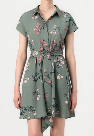 Bloemenjurk van groene stof met korte mouwen, kraag, gestrikt in de taille, met roze en donkere bloemenpatronen. Asymmetrische zoom.