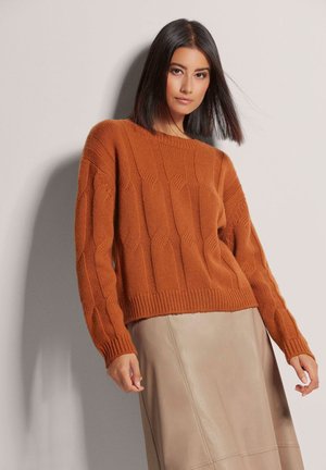 Roest-oranje gebreide sweater met een geribbelde textuur en verlaagde schouders, gecombineerd met een beige leren rok. De stof lijkt chunky en warm.