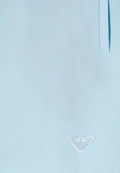 Ανοιχτό μπλε ύφασμα με ένα μικρό, ραμμένο λογότυπο αετού σε λευκό και μπλε κοντά στο κάτω μέρος και κάθετη ραφή στη δεξιά πλευρά.