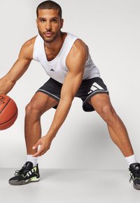 Top desportivo branco, calções pretos com bordas brancas, sapatilhas pretas e verdes. A segurar uma bola de basquete numa pose dinâmica e desportiva.