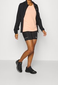 Giacca da corsa nera, top pesca, pantaloni corti neri con accenti bianchi e scarpe da atletica nere. Tessuti lisci e design aderente.