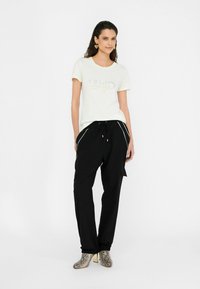 T-shirt crème à manches courtes avec lettering métallique, associé à un pantalon noir à coupe ample avec poches latérales et rayures blanches. Talons texturés à motif serpent.