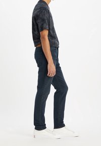 Hombre con camiseta negra con tintes anudados, jeans azul oscuro y zapatillas blancas, de pie frente a un fondo blanco liso.