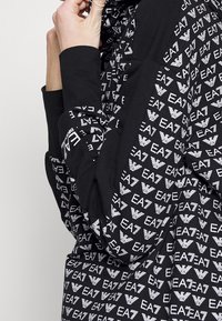 Czarna bluza z kapturem z wzorem białych logo EA7, z długimi rękawami i luźnym krojem. Materiał wydaje się miękki i teksturowany.