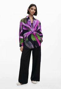 Chemise à manches longues en soie, présentant un imprimé floral audacieux en tons violets et verts sur un fond sombre. Associée à un pantalon large noir.