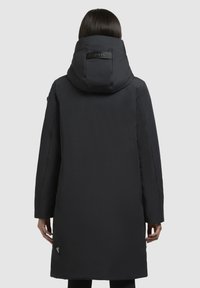 Manteau noir long avec un col haut, texture lisse et fermeture éclair dissimulée. Présente un label de marque dans le dos et une poche latérale.