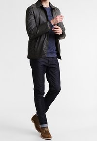 Uomo che sistema la manica indossando una giacca verde scuro, un maglione blu navy, jeans scuri e stivali in camoscio marrone, in piedi su uno sfondo bianco.