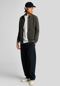 Giacca zip-up verde scuro, t-shirt bianca, pantaloni neri larghi e sneaker beige. Cappellino nero, branding minimale sulla giacca.