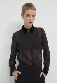Schwarze durchsichtige Bluse mit einem Samtkragen und -ärmeln, ausgestattet mit einer Knopfleiste und transparentem Stoff, kombiniert mit strukturierten schwarzen Hosen.