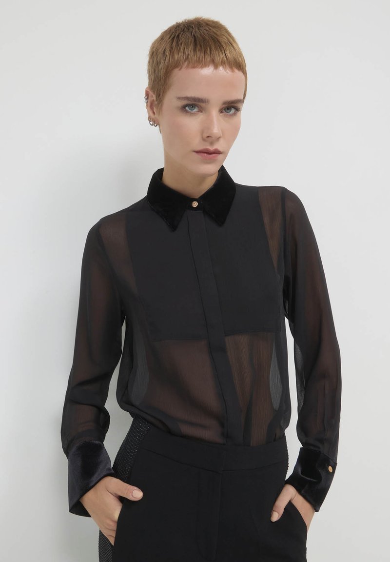 Schwarze durchsichtige Bluse mit einem Samtkragen und -ärmeln, ausgestattet mit einer Knopfleiste und transparentem Stoff, kombiniert mit strukturierten schwarzen Hosen.