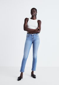 Schlanke blaue Jeans mit Ausbleichungen von dunkel nach hell, mit einer mittelhohen Taille und einem rohen Saum. Kombiniert mit einem weißen ärmellosen Oberteil und schwarzen Ballerinas.