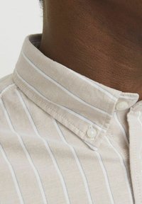 Lichtbeige shirt met dunne witte en grijze strepen. Heeft een button-down kraag en witte knoopsluitingen, en toont een gestructureerde stof.