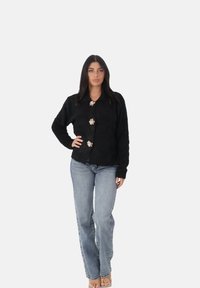 Cardigan nero peloso con colletto, tre bottoni floreali, indossato sopra jeans in denim azzurro chiaro. Tessuto morbido e vestibilità rilassata.