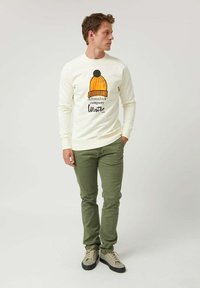 Sudadera crema con un gráfico de temática invernal, que presenta un gorro de punto amarillo con un pompón. Combinada con pantalones verdes y zapatillas beige.
