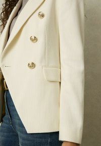 Svetločistý vzorovaný blejzer s tromi zlatými dekoračnými gombíkmi a náprsníkom s klopou, nosený cez sivý top a modré džínsy.